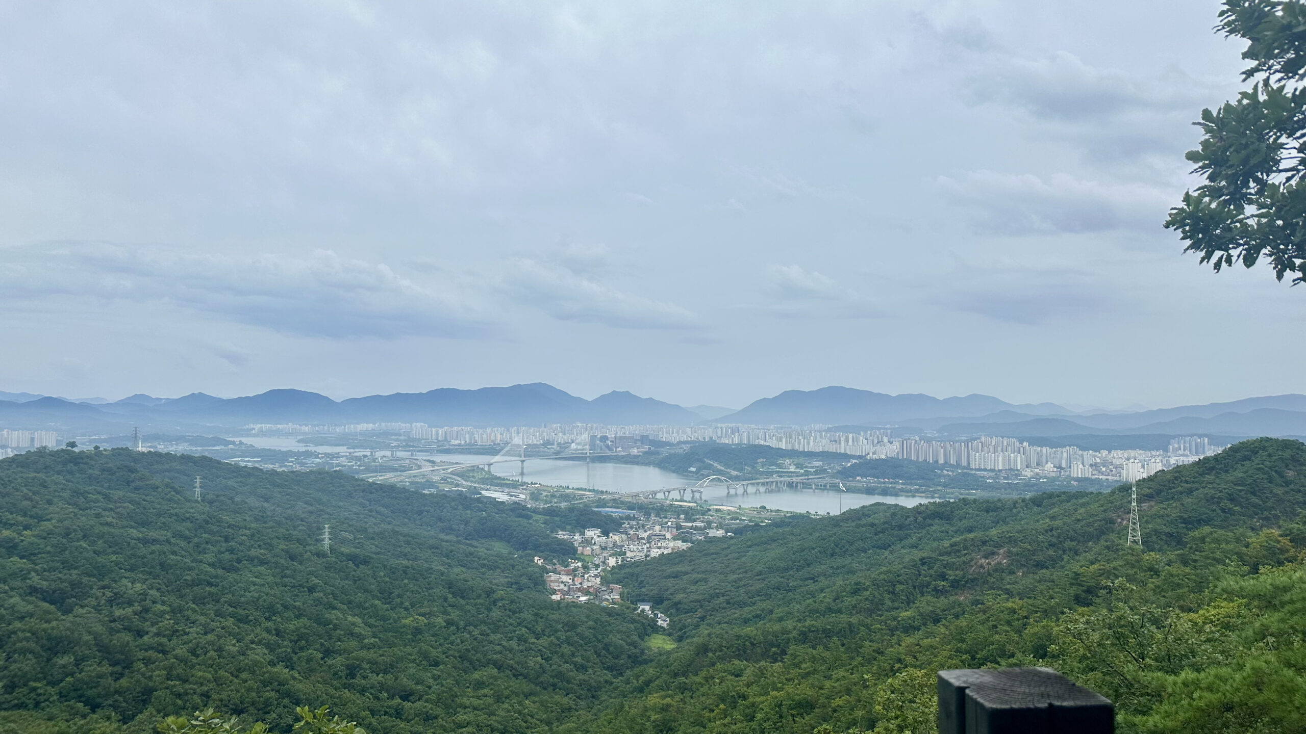 A view of Seoul from a lookout point on a mountainside. The silver colors of the city nestled in the valley is contrasted by the clear blue sky above, the dark colors of the mountains beyond the city, and the deep greens of the forest of the mountainside.