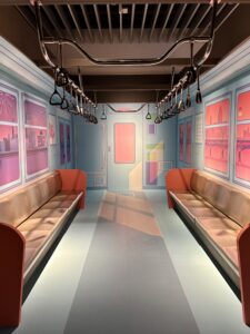 A life size depiction of a subway is taking up a whole room. There are seats on which someone could sit and two rails hang from the ceiling with individual handle grips like would be found on a real subway car.