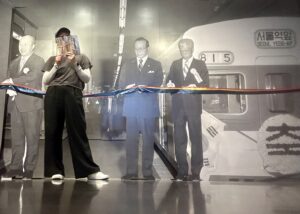 A black and white photo covers an entire wall. It shows a subway with three men standing on the platform. A ribbon hangs from the wall in their hands depicting a ribbon cutting. Gardner stands between the men in the picture with a program obstructing their face.