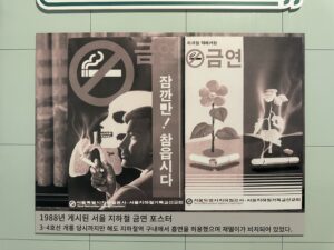 Two posters hang side by side. The poster on the left shows a no smoking sign with a skull and a the silhouette of a person smoking. The poster on the right has a flower on one side with a broken cigarette under it and a burned flower on the other with a lit cigarette under it. The advertisements are written in Korean.